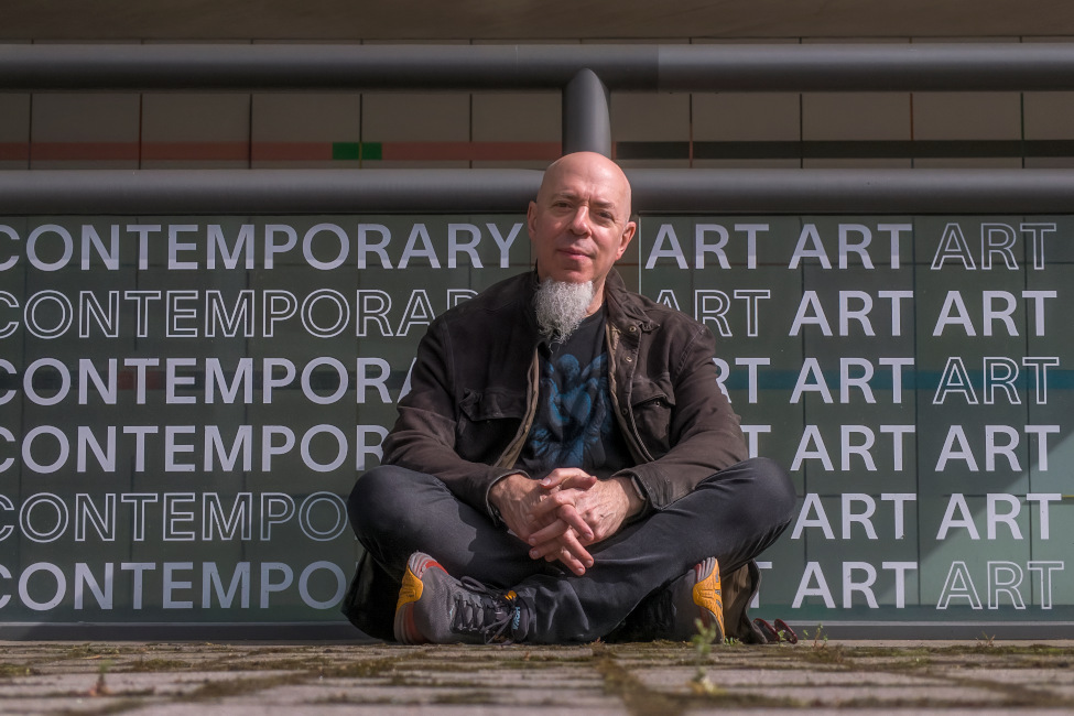 Der Künstler im Schneidersitz und mit weißem Kinnbart vor einer Wand mit der mehrfachen Aufschrift "Contemporary Art"