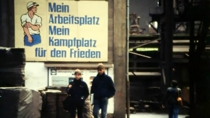 In einer ansonsten dunklen Werkhalle ist das erleuchtete Transparant mit einem Arbeier, der sich die Ärmel hochkrempelt, und der Aufschrift "Mein Arbeitsplatz Mein Kampfplatz für den Frieden" zu sehen.