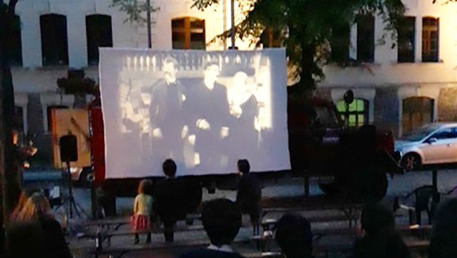 Auf dem Markt sitzen Menschen auf Holzbänken und verfolgen einen Film, der auf eine mobile Leinwand projeziert wird.