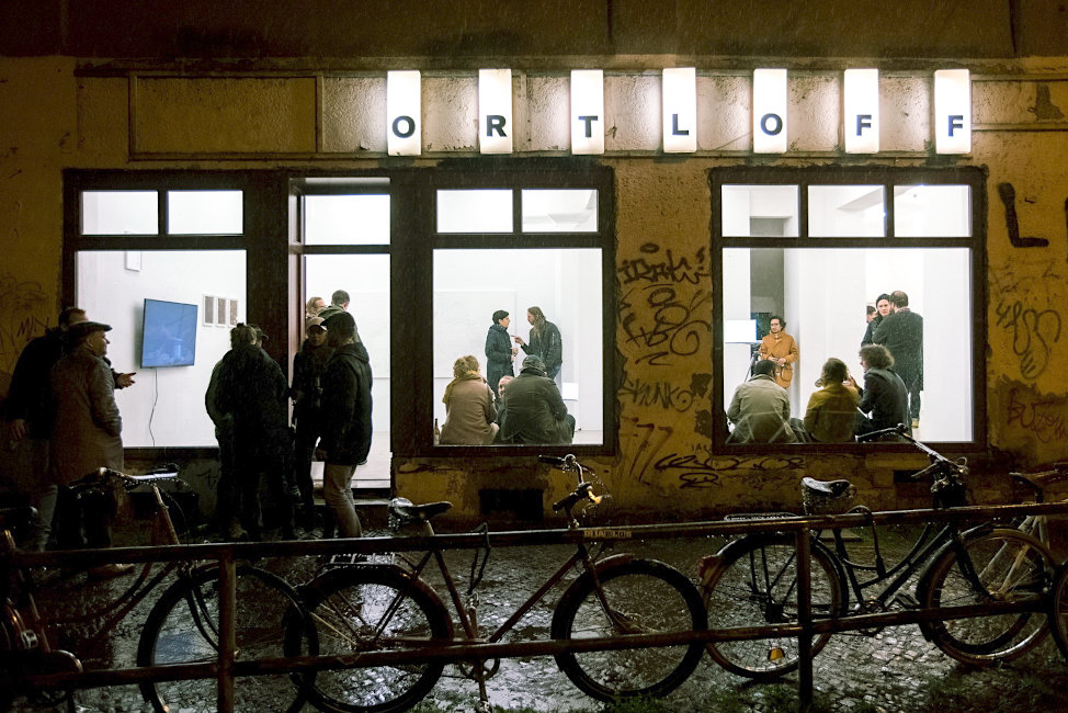 Ein Foto zeigt die hellerleuchteten großen Schaufenster einer Galerie mit einigen Besucherinnen und Besuchern darin und davir