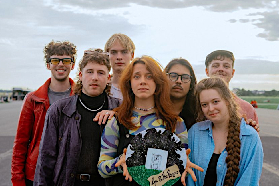 Gruppenfoto der ernst blickenden Band auf einem weiten betonierten Platz unter einem wolkenverhangenen Himmel