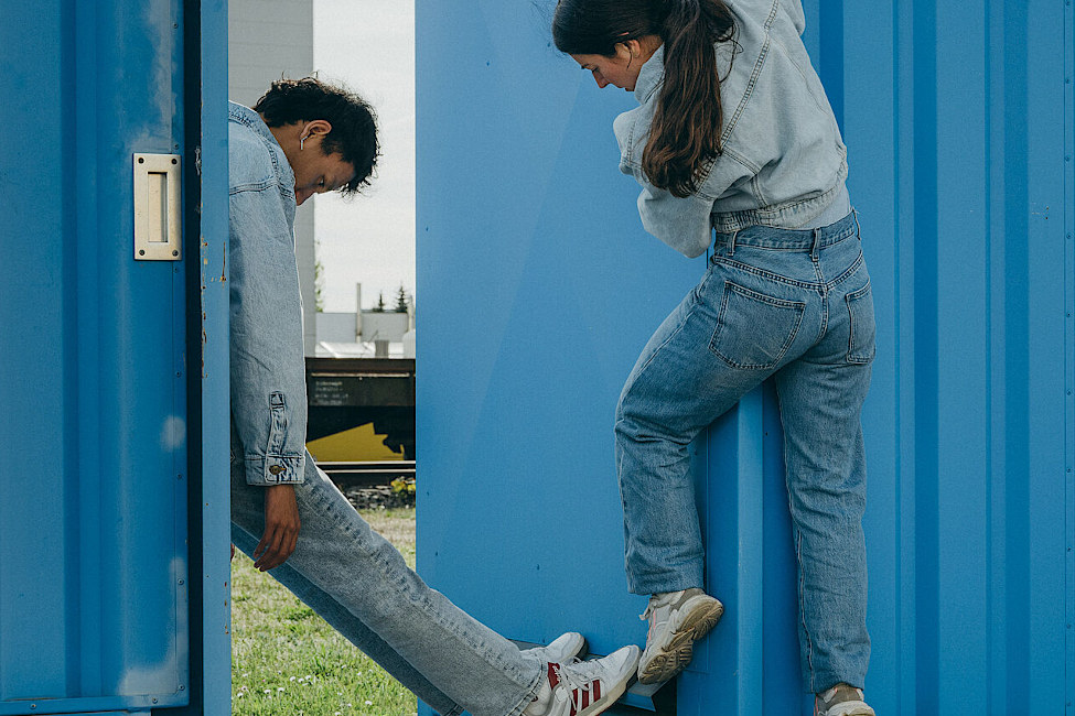 Ein junger Mann und eine junge Frau in Jeanskleidung klettern zwischen zwei blauen Containern