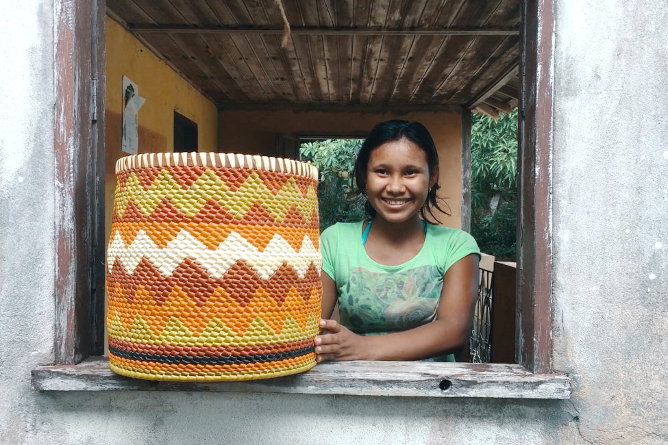 Eine junge Frau ist am Fenster eines einfachen Landhauses mit einer kunsvoll geflochtenen Flechtarbeit zu sehen.