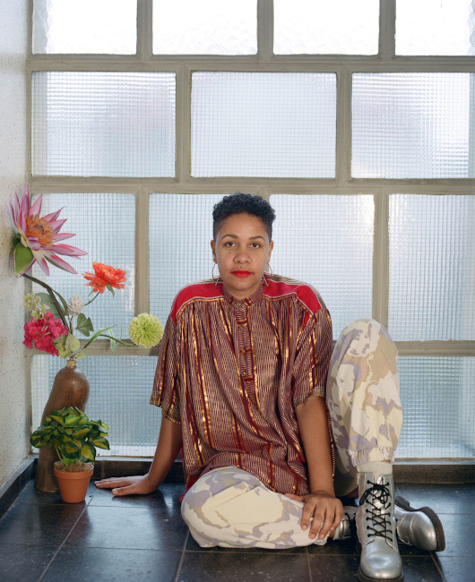 Die Künstlerin sitzt in einem seidig glänzenden Oberteil, Hosen mit Tarnmuster und Springerstiefeln auf dem Fliesenboden eines Treppenhauses.