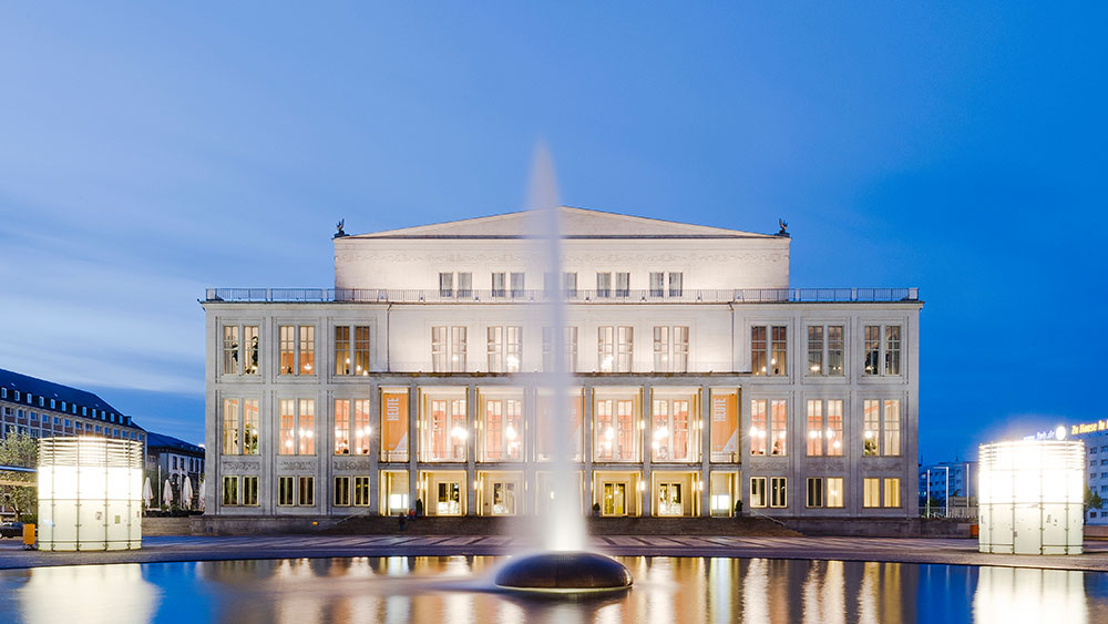 Das Opernhaus ist in der Dämmerung beleuchtet und davor spiegelt sich eine Fontaine im Wasserbecken auf dem Augustusplatz.
