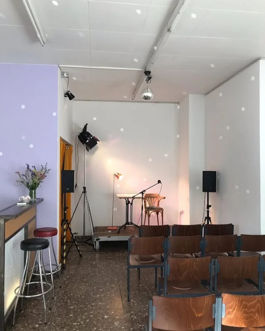 In einem schmalen Raum stehen vor einer kleinen Bühne mit Tisch und Stuhl Reihen von Holzstühlen.