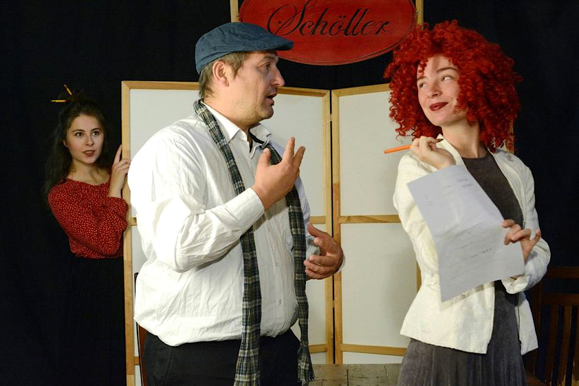 Ein Mann spricht zu einer Frau, die einen Stift und ein Blatt Papier in den Händen hält. Im Hintergrund ist eine junge Frau zu sehen, die die beiden beobachtet.