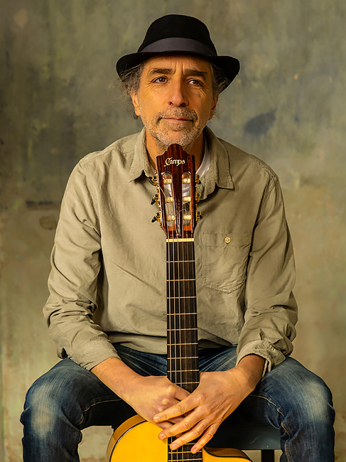 Der Künstler sitzt, mit seiner Gitarre vor sich, auf einem Stuhl und schaut verschmitzt in die Kamera