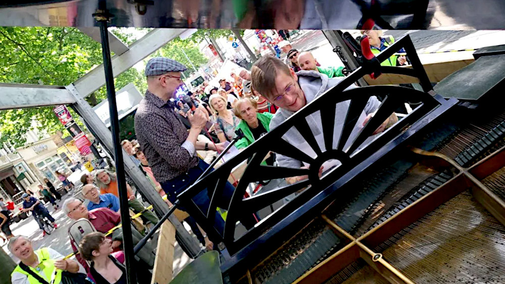 Auf der Ladefläche eines Fahrzeuges spielt ein Pianist auf einem Flügel, gefolgt von einem Protestzug.