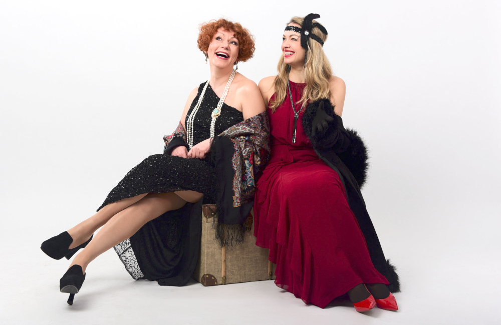 Die beiden Musikerinnen Lora Kostina und Katharina Eirich sitzen in Abendgarderobe lachend auf einem Koffer.