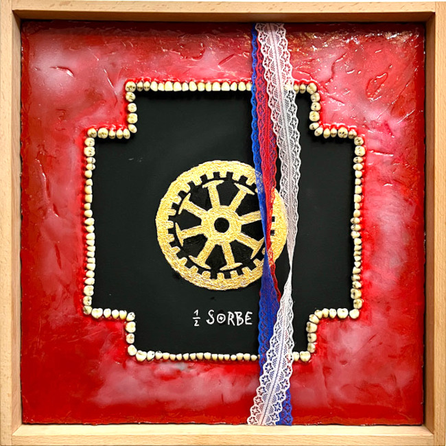 In einem quadratischen Holzrahem befindet sich ein schwarzes Kreuz mit einem goldenen Rad und der Aufschrift "1/2 SORBE".