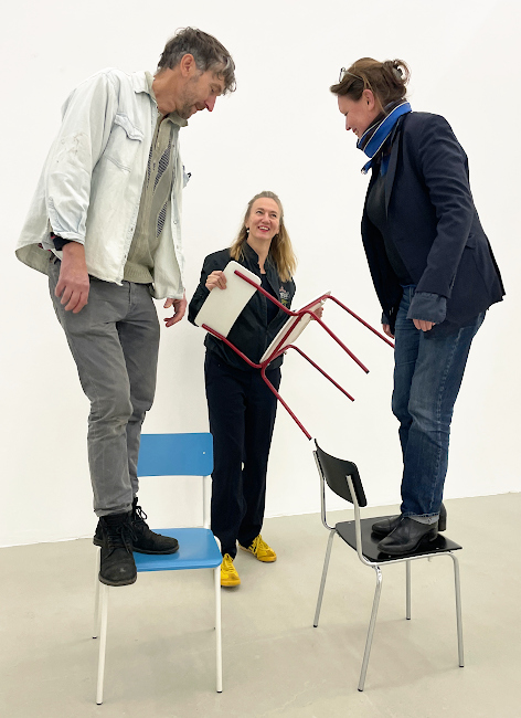 Foto der drei KünstlerInnen, die dabei sind, sich in einem leeren Raum auf Sthle zu stellen