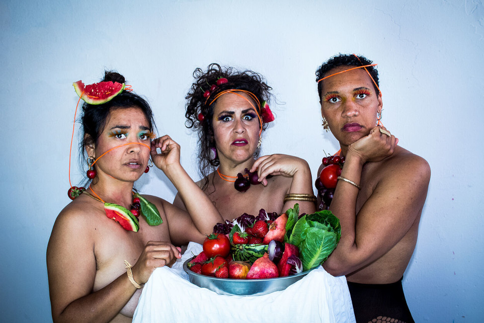 Drei Frauen mit nackten Oberkörpern stehen an einem hohen Tisch mit einer Schale voll Obst und Gemüse und schauen leicht angewiedert in die Kamera. Auf den Köpfen und um den Hals tragen sie ebenfalls Obst und Gemüse.