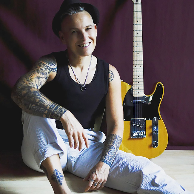Die Musikerin sitzt lächelnd vor ihrer Gitarre, die an der Wand lehnt, auf dem Boden.