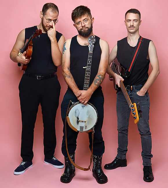 Gruppenfoto des skeptisch blickenden Trios mit seinen Instrumenten vor einem zartrosa Hintergrund