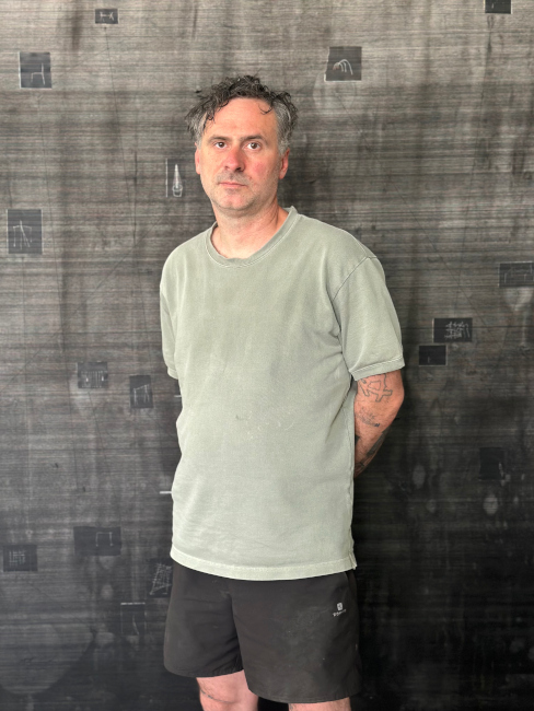 Der Künstler steht in T-Shiert und kurzer Hose vor einer Wand mit Texten und Fotografien.