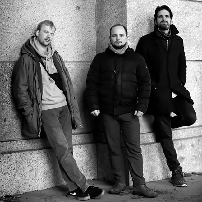 Die drei Musiker in Winterjacken lehnen sich an eine Wand.