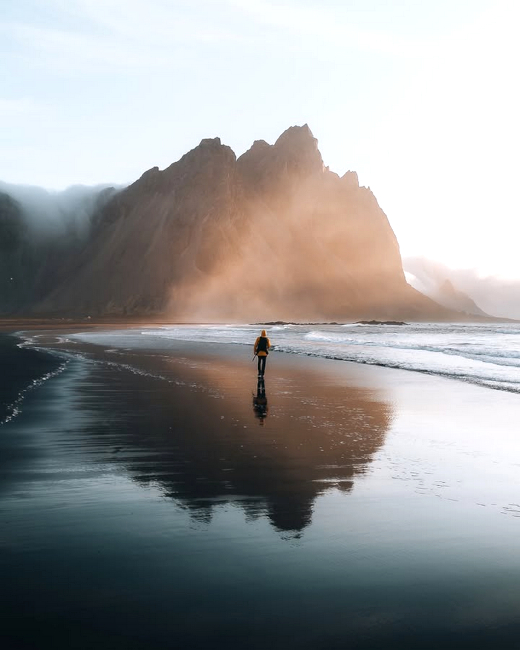 An einer Küste spiegelt sich ein Felsen im flachen Wasser, durch das eine Person läuft.