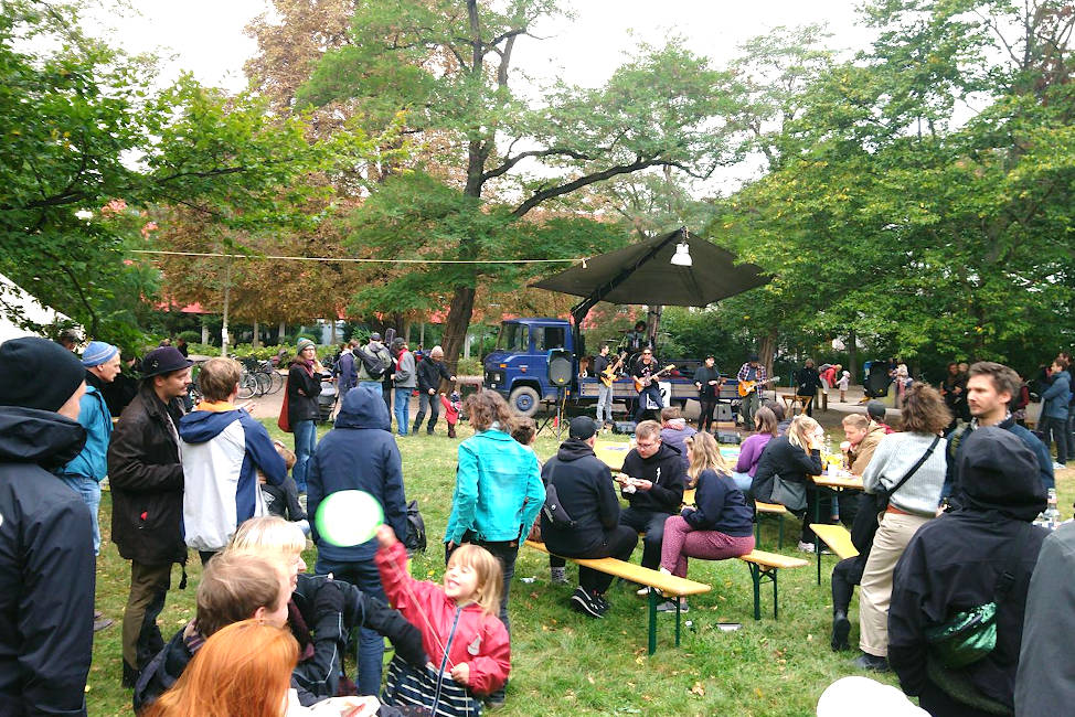 Auf einer großen Wiese stehen viele Menschen oder sitzen an Bierbänken und verfolgen ein Konzert auf einer kleinen Bühne.